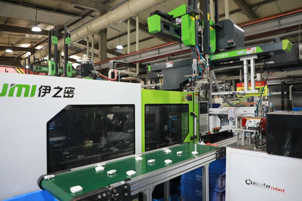 A robotic arm placing freshly moulded white plastic parts onto a conveyor next to an injection moulding machine