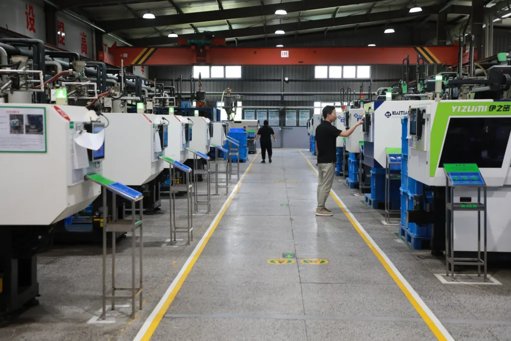 A wide row of injection moulding machines on the factory floor with two staff reviewing a machine control panel