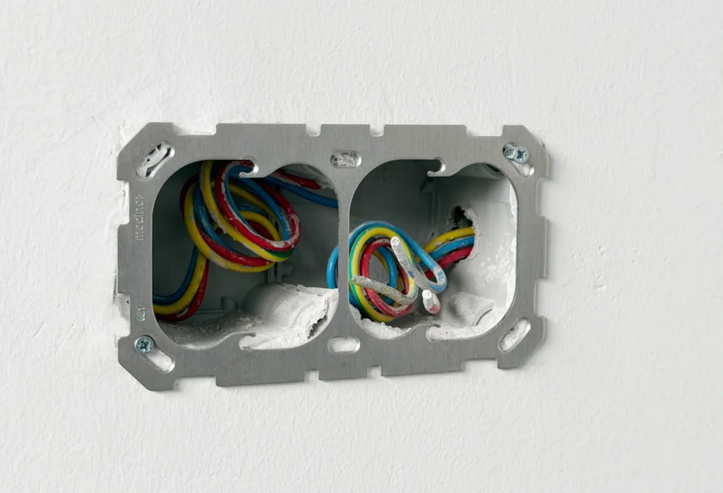 A metal double mounting frame fitted across two round installation boxes recessed in a plastered wall, with wiring visible through both openings