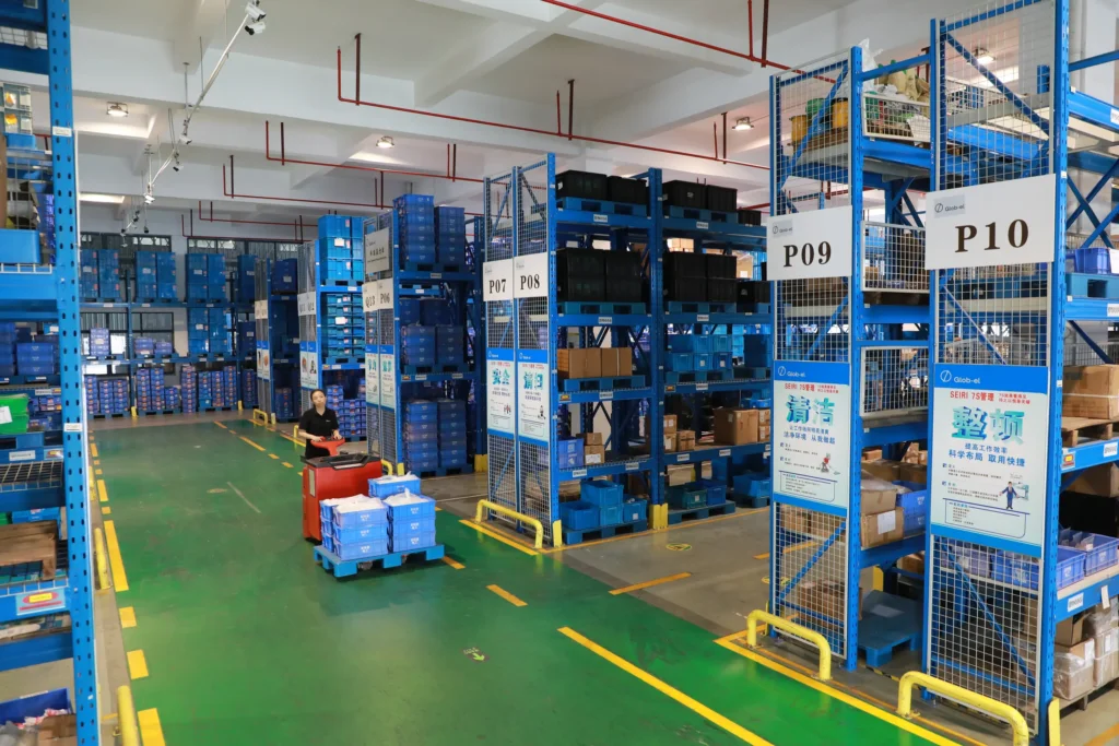 Organised warehouse racking with labelled storage bays and 5S lean management signage, a pallet truck moving stock